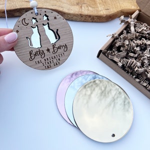 May include: A round wooden ornament with two cats cut out and the text "Betty & Barry The Brightest Stars In The Sky". The ornament is hanging from a string with a wooden bead. There are four round mirrors in the background.