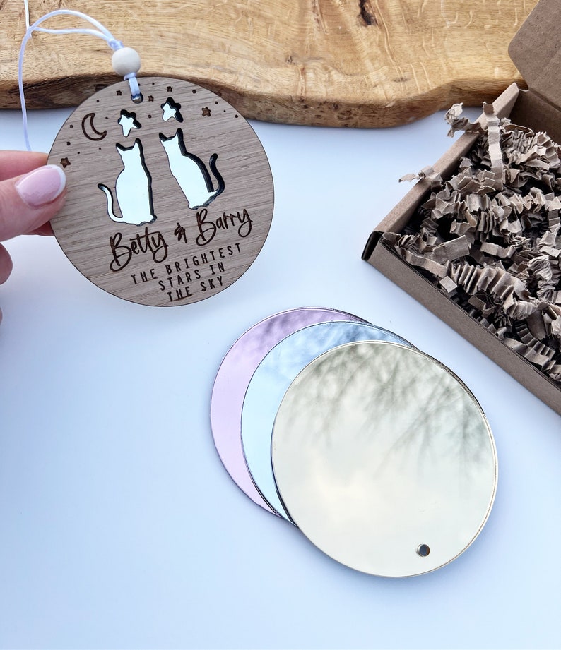 May include: A round wooden ornament with two cats cut out and the text "Betty & Barry The Brightest Stars In The Sky". The ornament is hanging from a string with a wooden bead. There are four round mirrors in the background.