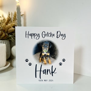 May include: A white card with the text "Happy Gotcha Day" and a photo of a black and tan puppy. The puppy is sitting on a concrete surface. The card also has the text "Hank" and "19th May 2024".