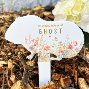 May include: White memorial garden stake shaped like a guinea pig with a floral pattern and the text "In loving memory of Ghost" in gold lettering.