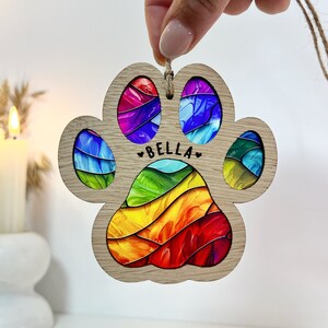 Peut inclure: Ornement en forme de patte d'animal avec un motif arc-en-ciel et le nom "BELLA" au centre. L'ornement est en bois et poss&egrave;de une ficelle pour l'accrocher.