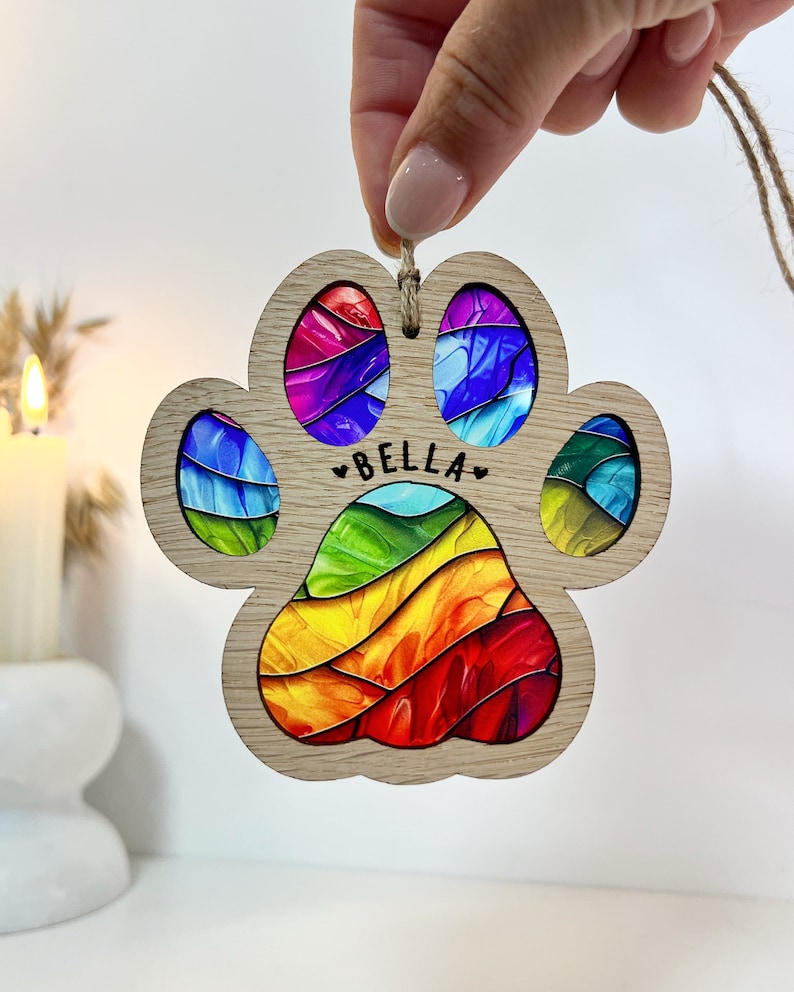 Peut inclure: Ornement en forme de patte d'animal avec un motif arc-en-ciel et le nom "BELLA" au centre. L'ornement est en bois et poss&egrave;de une ficelle pour l'accrocher.