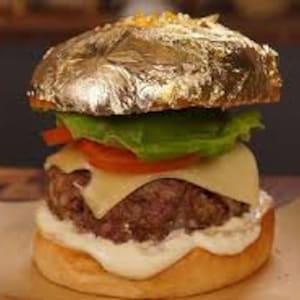 May include: A luxurious burger with a gold leaf bun, lettuce, tomato, cheese, and a beef patty. The burger is presented on white paper, with a blurred background.