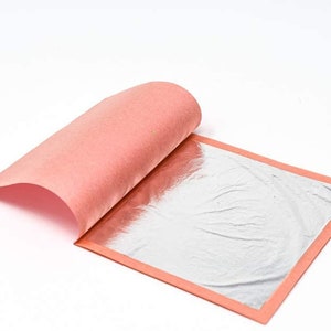 May include: A sheet of silver leaf with a pink backing, partially peeled back to reveal the shiny surface. The silver leaf is rectangular with a thin, orange-red border. The background is plain white.