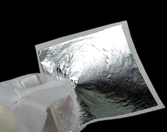 May include: A square sheet of thin, reflective silver leaf material with a crinkled, metallic texture. The silver foil is attached to a white paper backing, showcasing a highly polished, mirror-like surface that catches light across its uneven, delicate folds.