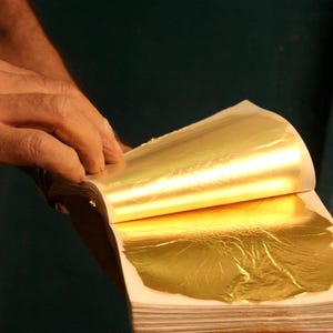 May include: A booklet of thin, metallic gold leaf sheets with a reflective, crinkled surface texture. The pages are separated by white paper layers, and the gold material appears delicate, lightweight, and highly polished with a bright yellow hue.