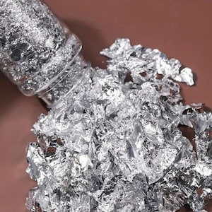 May include: A clear plastic bottle tilted downward, spilling out a pile of thin, crinkled, metallic silver foil flakes. The reflective material has an irregular, crumpled texture and a bright, shiny finish that catches the light across its surface.