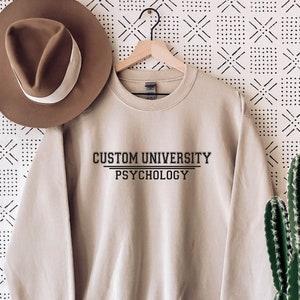 Könnte beinhalten: Ein beiger Pullover mit dem Aufdruck "CUSTOM UNIVERSITY PSYCHOLOGY" auf der Vorderseite.