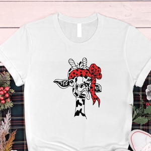 May include: White crew neck t-shirt with a black and white giraffe design. The giraffe wears a red polka-dot bandana and a red bow. The giraffe's tongue is visible. The shirt is made of a soft material.