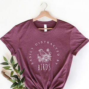 May include: A maroon t-shirt with a white graphic design that says "Easily Distracted By Birds" with a stylized bird and flowers.