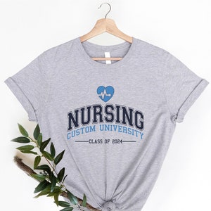May include: Gray t-shirt with a blue heart and a heartbeat graphic. The text "Nursing Custom University - Class of 2024" is printed on the shirt.