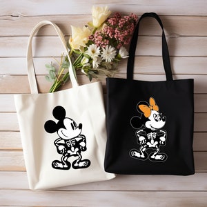 May include: Two white canvas tote bags with black handles. One bag features a black and white illustration of Mickey Mouse as a skeleton. The other bag features a black and white illustration of Minnie Mouse as a skeleton with an orange bow.