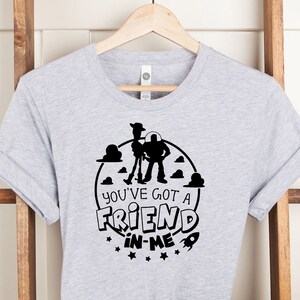 Może przedstawiać: Szara koszulka z czarną sylwetką Woody'ego i Buzz Lightyeara z Toy Story. Projekt zawiera koło z chmurami i gwiazdami oraz tekst "You've got a friend in me".