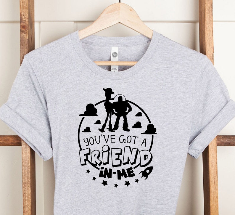 Może przedstawiać: Szara koszulka z czarną sylwetką Woody'ego i Buzz Lightyeara z Toy Story. Projekt zawiera koło z chmurami i gwiazdami oraz tekst "You've got a friend in me".