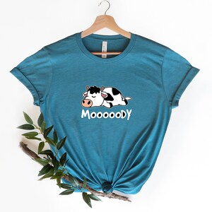 以下が含まれることがあります： 漫画の牛が寝ている、ターコイズブルーのTシャツ。前面に「MOOOOODY」の文字がプリントされています。
