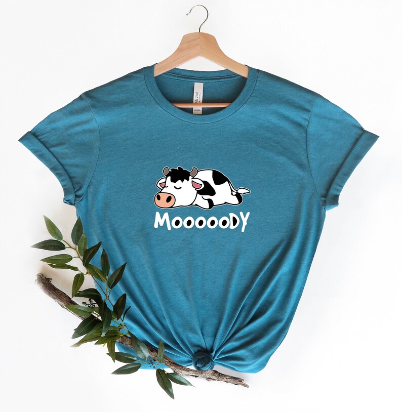 以下が含まれることがあります： 漫画の牛が寝ている、ターコイズブルーのTシャツ。前面に「MOOOOODY」の文字がプリントされています。