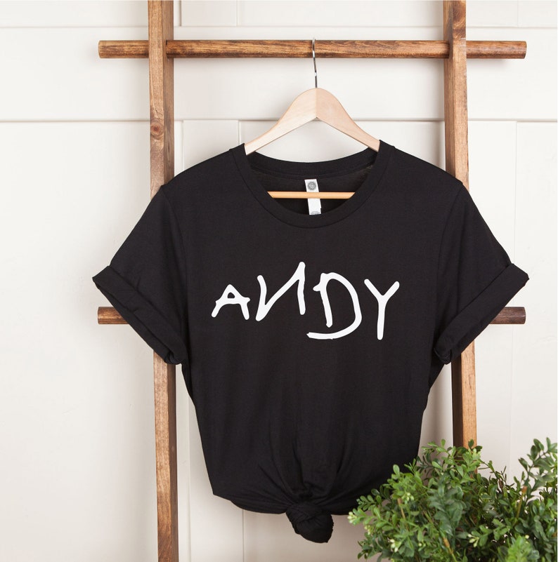Może przedstawiać: Czarny T-shirt z białym napisem "ANDY" w stylu odręcznym. Koszulka wisi na drewnianej drabinie.