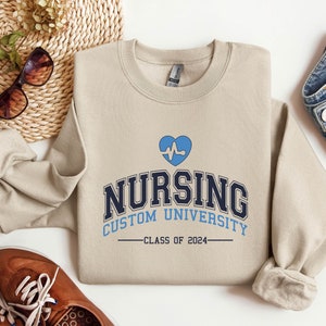Puede incluir: Una sudadera de color beige con un gráfico de corazón azul y el texto "Nursing Custom University - Class of 2024".