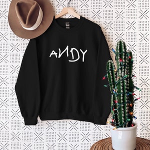 Może przedstawiać: Czarny sweter z napisem "ANDY" w kolorze białym na przodzie. Sweter wisi na drewnianym wieszaku na tle białego tła z czarno-białym geometrycznym wzorem.