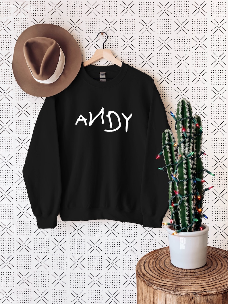 Może przedstawiać: Czarny sweter z napisem "ANDY" w kolorze białym na przodzie. Sweter wisi na drewnianym wieszaku na tle białego tła z czarno-białym geometrycznym wzorem.