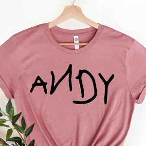 Może przedstawiać: R&oacute;żowy t-shirt w kolorze pudrowego r&oacute;żu z napisem "ANDY" napisanym czarnym kolorem, w dziecięcym stylu.