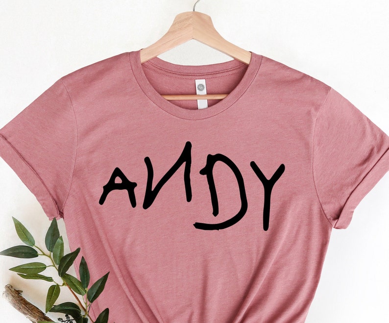 Może przedstawiać: R&oacute;żowy t-shirt w kolorze pudrowego r&oacute;żu z napisem "ANDY" napisanym czarnym kolorem, w dziecięcym stylu.