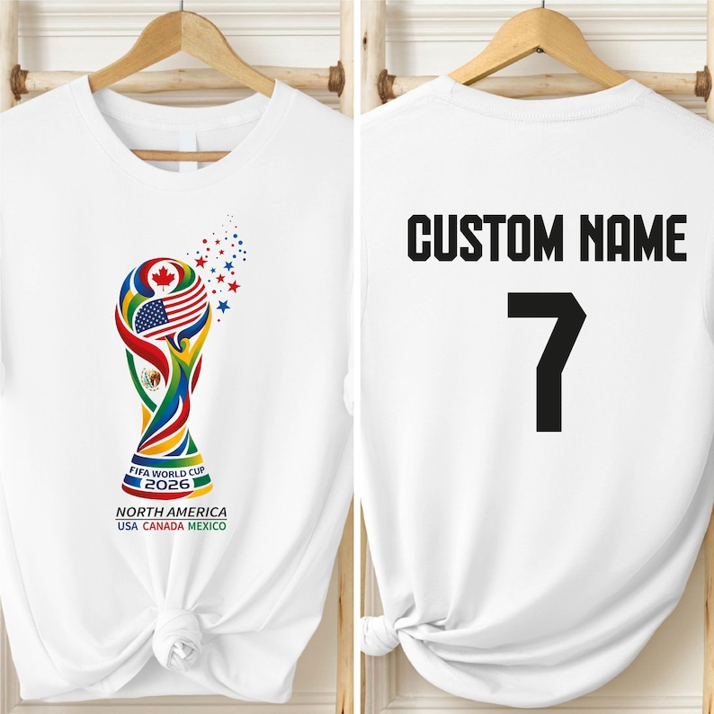 A white t-shirt with a colorful design featuring the FIFA World Cup trophy and the text "FIFA WORLD CUP NORTH AMERICA USA CANADA MEXICO" printed on it. The t-shirt also has the text "CUSTOM NAME" and the number "7" printed on the back.