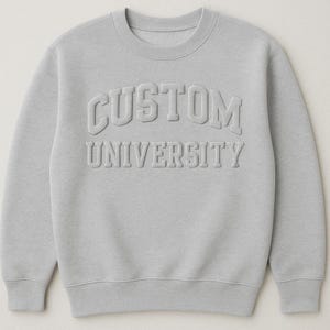 以下が含まれることがあります： 胸に「CUSTOM UNIVERSITY」の文字がエンボス加工された、明るいグレーのスウェットシャツ。クルーネックで長袖。文字は浮き上がった質感のあるフォントです。