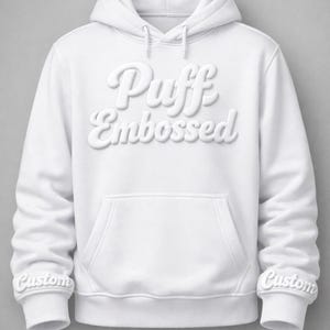 Puede incluir: Una sudadera con capucha blanca con un bolsillo delantero y una capucha con cordón. Las palabras "Puff Embossed" están en relieve en el pecho. Los puños de las mangas tienen la palabra "Custom".