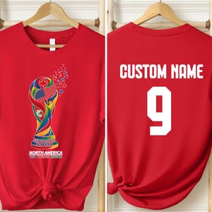 A red t-shirt with a colorful graphic of the FIFA World Cup trophy and the text "NORTH AMERICA USA CANADA MEXICO" printed on it. The t-shirt also has the text "CUSTOM NAME" and the number "9" printed on the back.