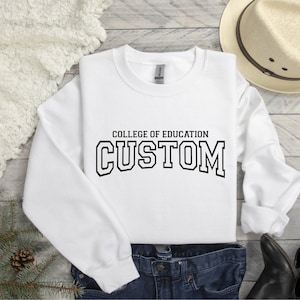 Könnte beinhalten: Weißer Pullover mit schwarzem Text, der "College of Education Custom" lautet.