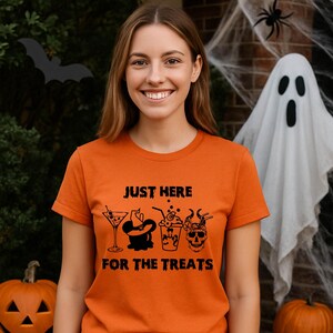 Puede incluir: Camiseta naranja con el texto "JUST HERE FOR THE TREATS" y gráficos temáticos de Halloween. El diseño incluye una copa de martini, un sombrero de bruja, una bebida y una calavera. Una mujer lleva la camiseta.