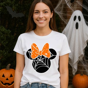 Pode incluir: Camiseta branca com um design da Minnie Mouse com tema de Halloween. O design inclui uma silhueta preta da cabeça da Minnie com uma teia de aranha e um laço laranja com detalhes de aranha. O fundo inclui decorações de Halloween.