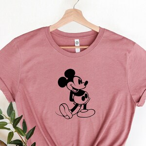 May include: A dusty rose t-shirt with a black silhouette of Mickey Mouse. Mickey is facing right and has his right hand on his hip.