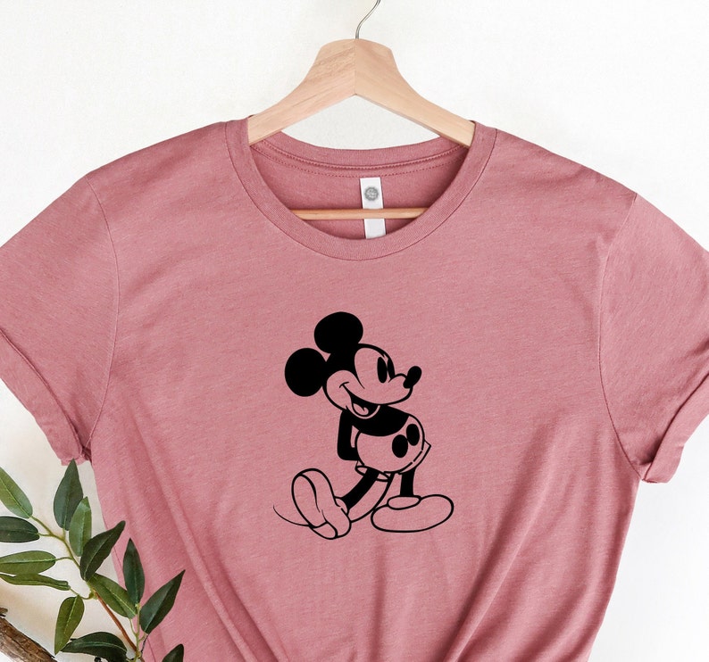 May include: A dusty rose t-shirt with a black silhouette of Mickey Mouse. Mickey is facing right and has his right hand on his hip.