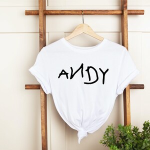 Może przedstawiać: Biała koszulka z napisem "ANDY" napisanym czarnym kolorem, stylem pisma odręcznego.