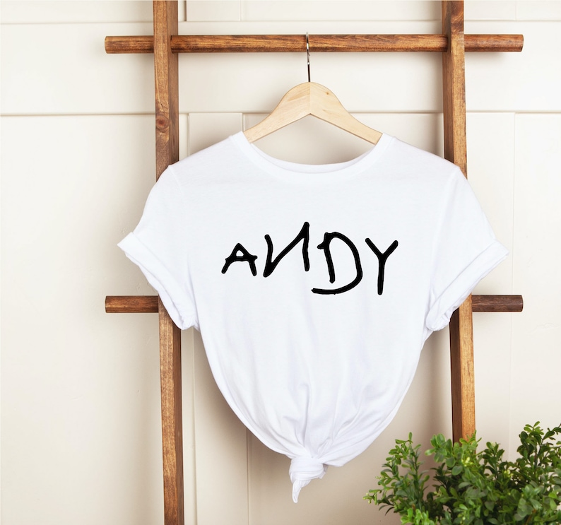 Może przedstawiać: Biała koszulka z napisem "ANDY" napisanym czarnym kolorem, stylem pisma odręcznego.