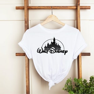 K&ouml;nnte beinhalten: Wei&szlig;es T-Shirt mit einer schwarzen Silhouette eines Schlosses in einem Kreis und den Worten "Walt Disney" in einer stilisierten Schriftart. Das T-Shirt h&auml;ngt an einem Holzb&uuml;gel.