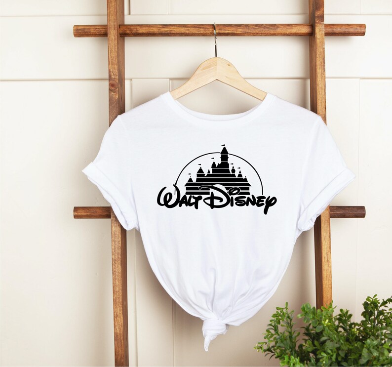 K&ouml;nnte beinhalten: Wei&szlig;es T-Shirt mit einer schwarzen Silhouette eines Schlosses in einem Kreis und den Worten "Walt Disney" in einer stilisierten Schriftart. Das T-Shirt h&auml;ngt an einem Holzb&uuml;gel.