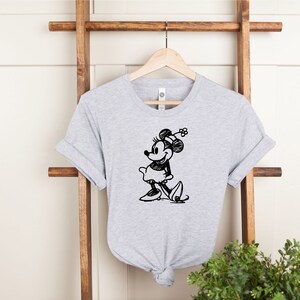 Pu&ograve; includere: Una maglietta grigia melange con uno schizzo in bianco e nero di Minnie Mouse sul davanti. Minnie Mouse indossa un vestito e delle scarpe e ha un fiocco sulla testa.
