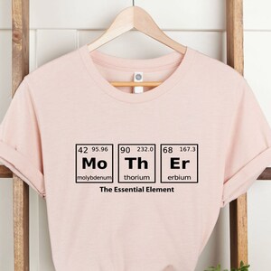 May include: A pink t-shirt featuring a graphic of three periodic table elements: Molybdenum (Mo), Thorium (Th), and Erbium (Er). The text "The Essential Element" is printed below the elements.