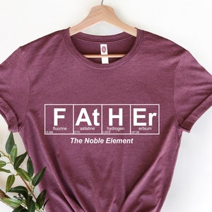 May include: Burgundy t-shirt with a white graphic of the word "FATHER" spelled out using the chemical symbols for the elements fluorine, astatine, hydrogen, and erbium. The text "The Noble Element" is printed below the graphic.