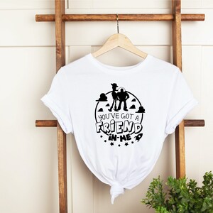 Może przedstawiać: Biała koszulka z czarnym nadrukiem Woody'ego i Buzza Astrala z Toy Story. Nadruk zawiera tekst "You've got a friend in me" i rakietę.