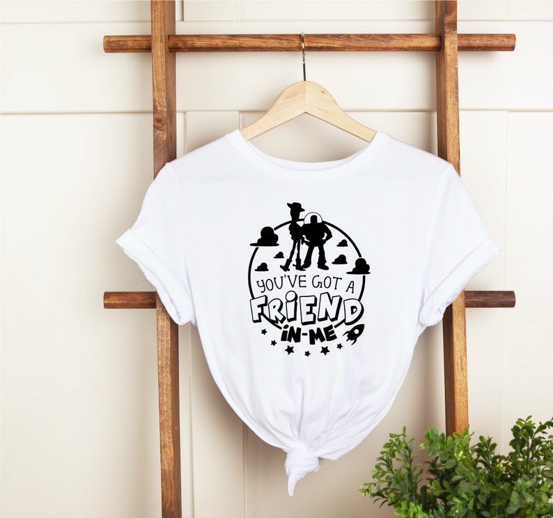 Może przedstawiać: Biała koszulka z czarnym nadrukiem Woody'ego i Buzza Astrala z Toy Story. Nadruk zawiera tekst "You've got a friend in me" i rakietę.