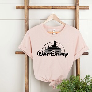 K&ouml;nnte beinhalten: Hellrosa T-Shirt mit schwarzer Grafik eines Schlosses in einem Halbkreis und dem Schriftzug "Walt Disney" darunter. Das T-Shirt h&auml;ngt an einem Holzkleiderb&uuml;gel.