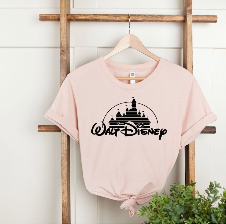 K&ouml;nnte beinhalten: Hellrosa T-Shirt mit schwarzer Grafik eines Schlosses in einem Halbkreis und dem Schriftzug "Walt Disney" darunter. Das T-Shirt h&auml;ngt an einem Holzkleiderb&uuml;gel.