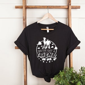Może przedstawiać: Czarny T-shirt z białym nadrukiem Woody'ego i Buzza Astrala z Toy Story. Nadruk zawiera tekst "You've got a friend in me" z gwiazdami i rakietą.
