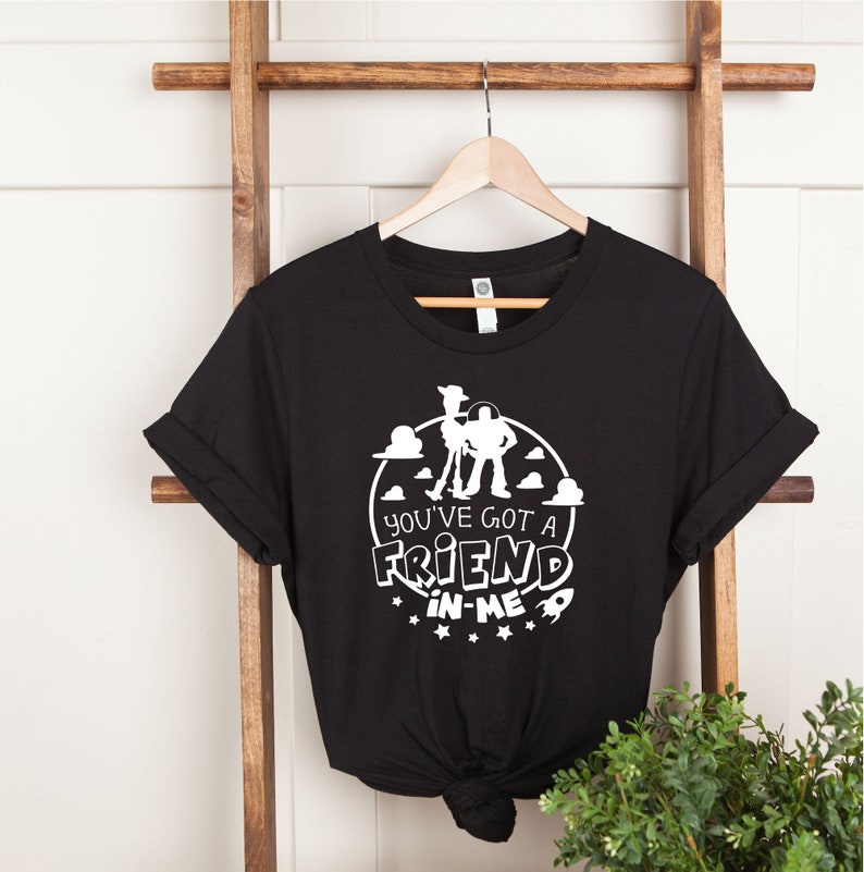 Może przedstawiać: Czarny T-shirt z białym nadrukiem Woody'ego i Buzza Astrala z Toy Story. Nadruk zawiera tekst "You've got a friend in me" z gwiazdami i rakietą.