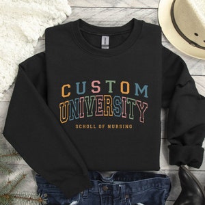 May include: Black sweatshirt with the text "CUSTOM UNIVERSITY" in rainbow colors and the text "SCHOLL OF NURSING" in white below.