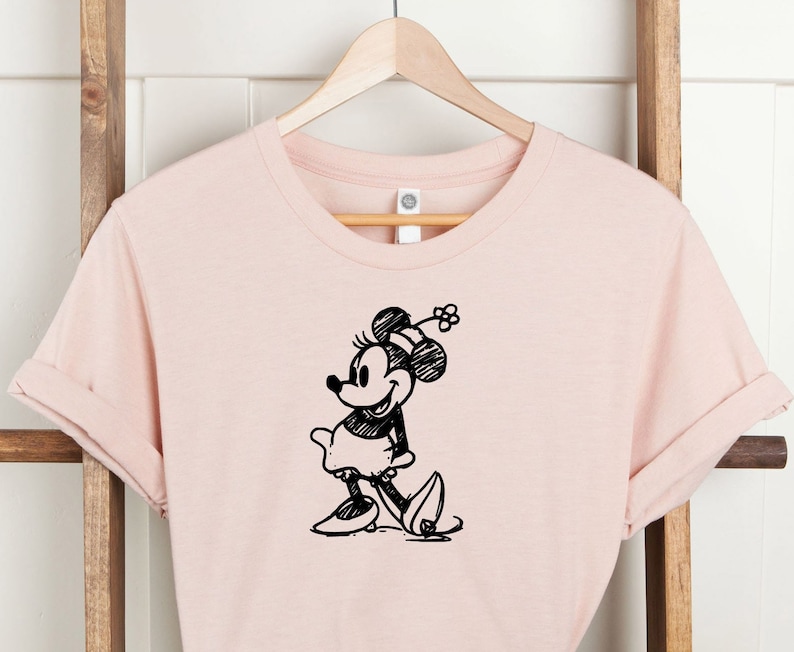 Pu&ograve; includere: Una maglietta rosa con uno schizzo in bianco e nero di Minnie Mouse sul davanti.